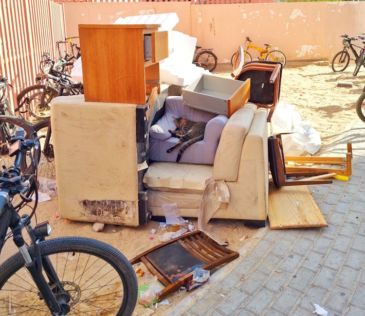 An assortment of discarded furniture and household items arranged on a pavement outdoors, including an old, heavily stained beige sofa with visible dirt and damage at its base, topped by a smaller purple armchair with a tabby cat lying across its cushions. Several other furniture pieces, such as a wooden cabinet, a small chest of drawers, and a broken wooden chair, are stacked haphazardly around the sofa. Some items are partially disassembled or tipped over, with pieces like a wooden plank, broken slats, and white packaging material scattered nearby. In the background, a low brick wall and gravel surface are visible, with bicycles leaning against the wall, indicating the scene is in a residential or communal outdoor space. The overall setting suggests a private waste clearance or alternative rubbish removal, handled by a professional service like Rubbish Clearance Wimbledon, to facilitate furniture disposal or site clearance for renovation or decluttering.