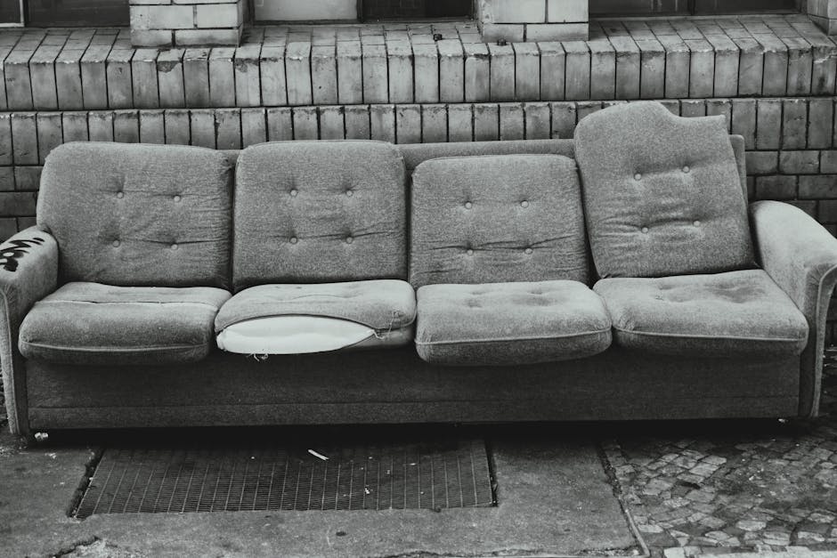 A worn-out, grey upholstered sofa with a textured fabric surface, positioned outdoors against a brick wall. The sofa features four seat cushions and four backrest cushions, each with fabric buttons, and shows visible signs of wear such as creases and slight sagging. One seat cushion has a tear revealing the white foam padding underneath. The sofa's armrests are rounded and covered in the same fabric, with the right armrest displaying some wear. It sits on a paved outdoor surface, with a small metal gutter grille directly beneath it. The brick wall behind is composed of rectangular bricks in a traditional pattern, with noticeable mortar joints, providing a neutral, urban background. The scene is lit with natural daylight, emphasizing the textures and details of the fabric and the surrounding environment. The image aligns with private waste handling and furniture clearance services, showcasing a typical piece of furniture prepared for disposal or collection by Rubbish Clearance Wimbledon, who specialise in rubbish removal services.
