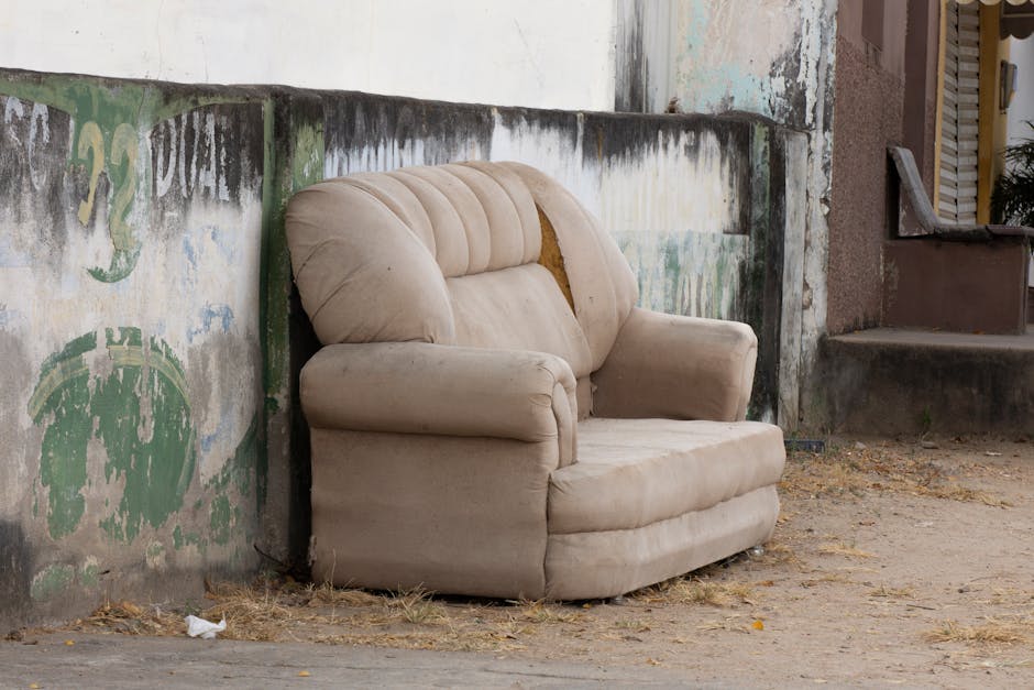 An assortment of discarded furniture and household items arranged on a pavement outdoors, including an old, heavily stained beige sofa with visible dirt and damage at its base, topped by a smaller purple armchair with a tabby cat lying across its cushions. Several other furniture pieces, such as a wooden cabinet, a small chest of drawers, and a broken wooden chair, are stacked haphazardly around the sofa. Some items are partially disassembled or tipped over, with pieces like a wooden plank, broken slats, and white packaging material scattered nearby. In the background, a low brick wall and gravel surface are visible, with bicycles leaning against the wall, indicating the scene is in a residential or communal outdoor space. The overall setting suggests a private waste clearance or alternative rubbish removal, handled by a professional service like Rubbish Clearance Wimbledon, to facilitate furniture disposal or site clearance for renovation or decluttering.