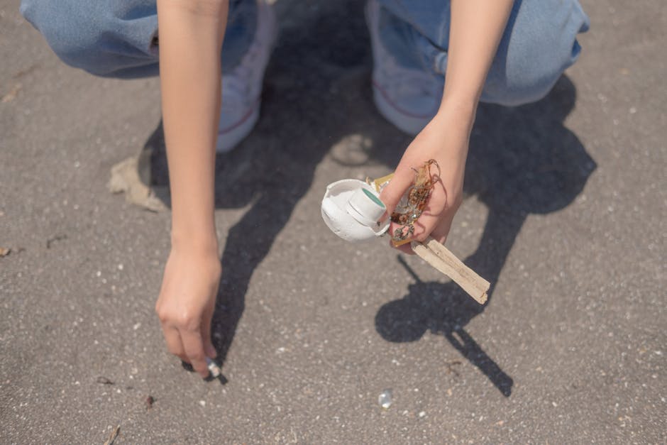 A person is crouching on a paved surface, holding a small collection of discarded rubbish in their right hand. The rubbish includes a white disposable coffee cup with a green logo, some tangled paper or cardboard debris, and a wooden stick or chopstick. The individual is wearing light blue jeans and white sneakers with pink accents. The background consists of the textured asphalt surface, and the shadows cast suggest a bright, sunny day. This scene reflects activities associated with littering, which may be subject to cleanup or waste management services like Rubbish Clearance Wimbledon. The setting appears outdoors on a driveway or footpath, with no other objects or environmental features visible.