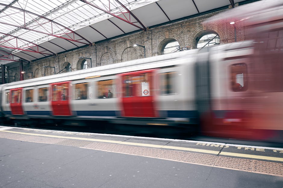 A London Underground train moving quickly through a station with an arching brick wall backdrop. The train has a white upper section, with red and blue stripes along the sides, and appears blurred due to motion. The platform in the foreground is surfaced with dark paving and features tactile paving strips near the edge for safety. The station's ceiling is constructed with metal beams and transparent panels, allowing natural light to illuminate the scene. Behind the train, the brick wall features several arched windows and black drainpipes affixed to the surface. The overall scene captures the sense of rapid commuter transit within an urban railway environment, relevant to private and independent waste removal or site clearance services often supporting busy infrastructure areas.