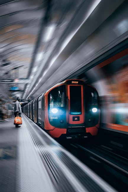 A modern urban underground train arriving or departing at a station, with a sleek, aerodynamic exterior painted in shades of blue and red. The train has illuminated front and side windows, and visible metal doors for passenger entry. The platform area features a blurred background indicating motion, with a passenger standing on the left side carrying an orange and black bag, dressed in casual clothing and looking toward the train. The station's ceiling and walls, made of reflective metallic surfaces, curve overhead, creating a dynamic sense of movement and spaciousness. The station lighting is bright, highlighting the train's smooth finish and the clean, organized environment typical of private or alternative waste handling facilities, such as those used by services like Rubbish Clearance Wimbledon, which can facilitate efficient rubbish collection or on-site clearance in dense urban areas. The overall scene emphasizes a structured, professional transportation environment that complements the company's focus on quick and reliable rubbish removal or disposal, aligning with the concept of alternative waste collection methods within busy city infrastructure.